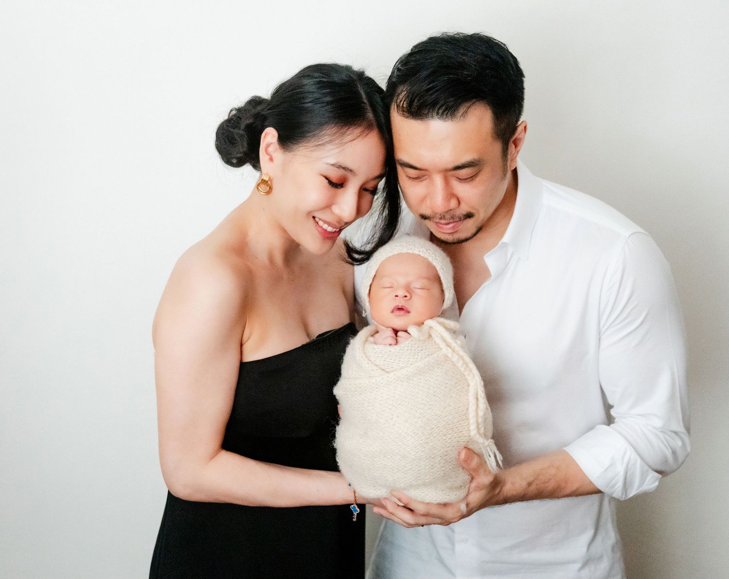 A couple in black and white outfits tenderly hold their swaddled newborn baby during a studio photo session.