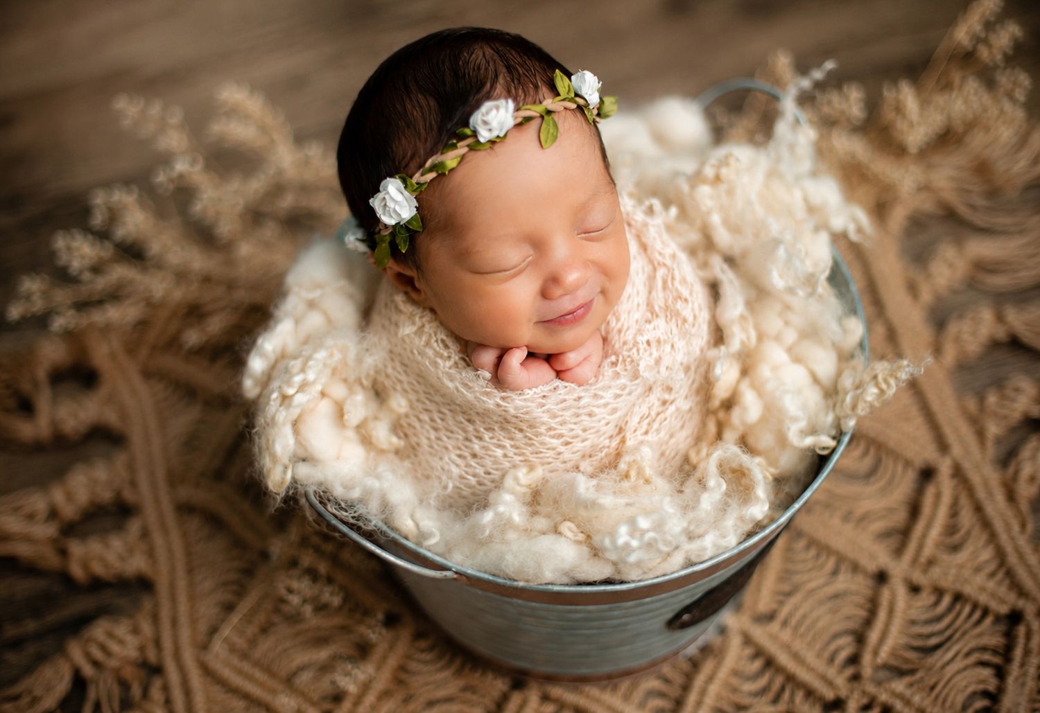 Smiling newborn baby wrapped in cream knit blanket, sitting in a metal bucket with a floral headband.