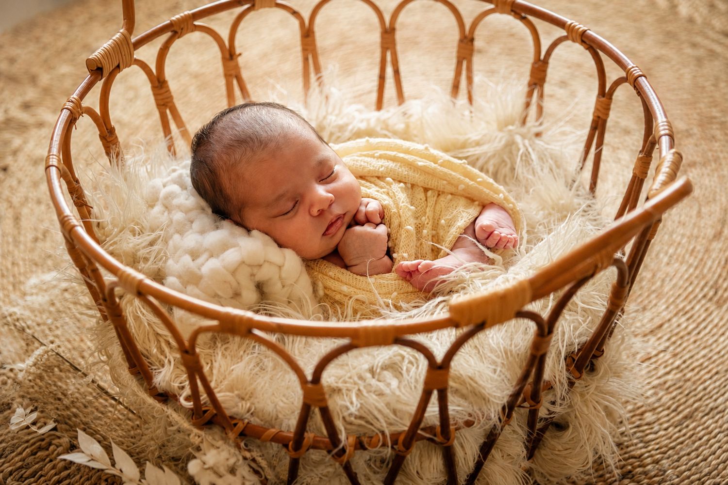 Newborn baby sleeping peacefully in a wicker basket lined with fluffy white fur, wrapped in a soft yellow blanket.