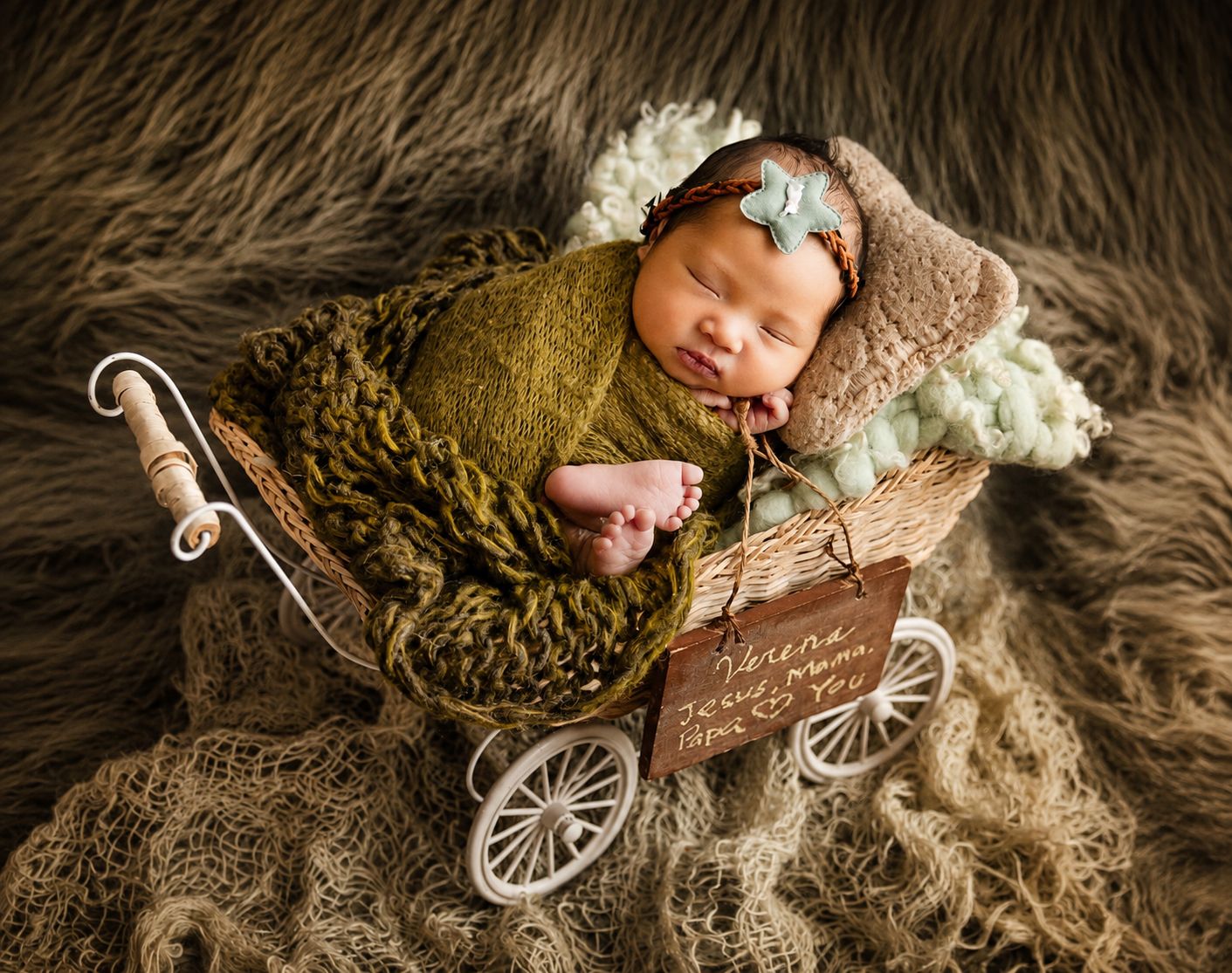 Newborn baby sleeping in a miniature vintage pram prop, wrapped in olive green knit, on a fluffy brown fur backdrop.