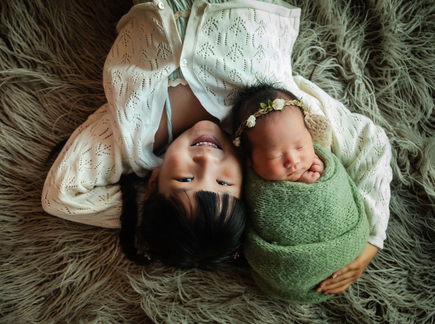 Mother and newborn baby lying together on a fluffy gray rug, infant swaddled in green wrap with floral headband.