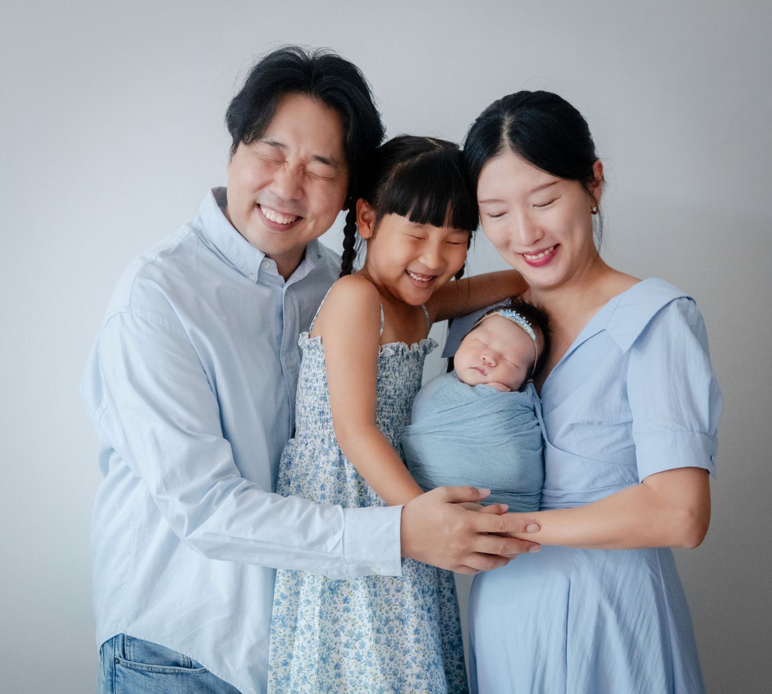 Happy Asian family of four posing together, parents smiling while mother holds swaddled newborn baby, with young daughter between them.