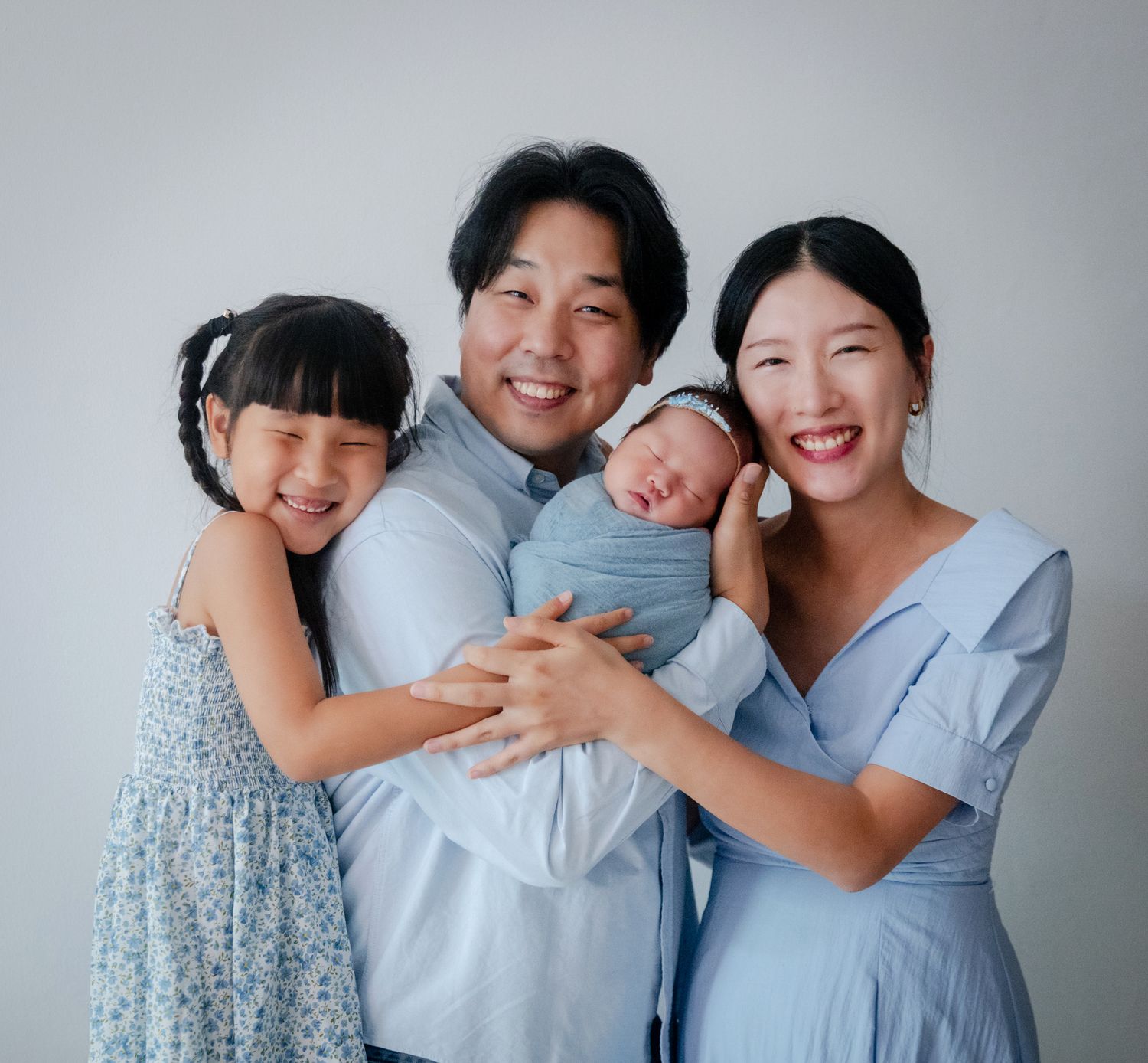 Happy Asian family of four posing together, parents holding newborn baby and young daughter smiling in blue outfits.