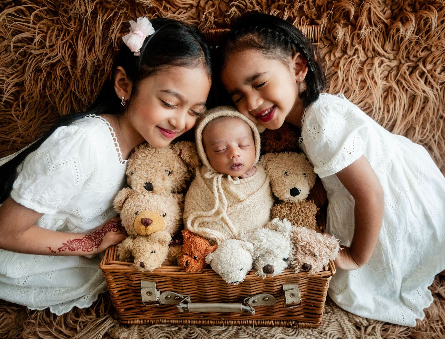 Two girls in white dresses smile beside a sleeping newborn wrapped in a basket with teddy bears.