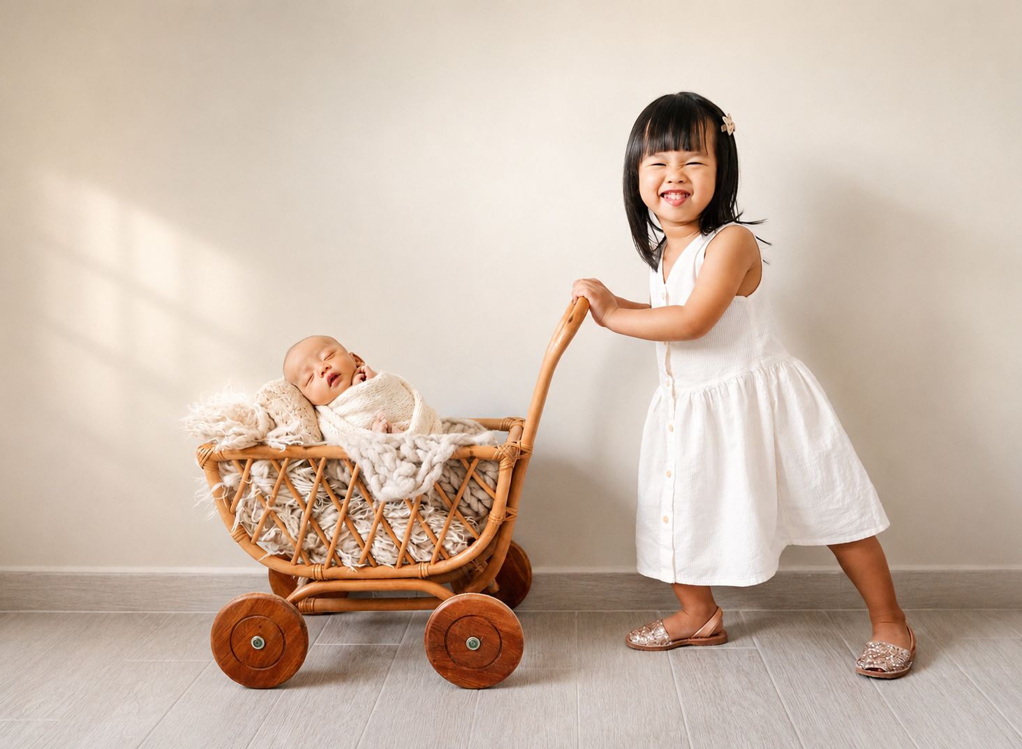 A smiling toddler girl in a white dress pushes a wicker toy pram with a sleeping newborn baby inside.