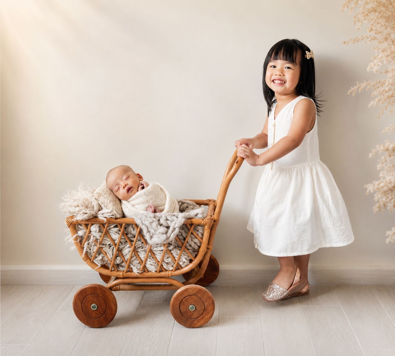 A smiling toddler girl in a white dress pushes a wicker pram with a sleeping newborn baby inside.