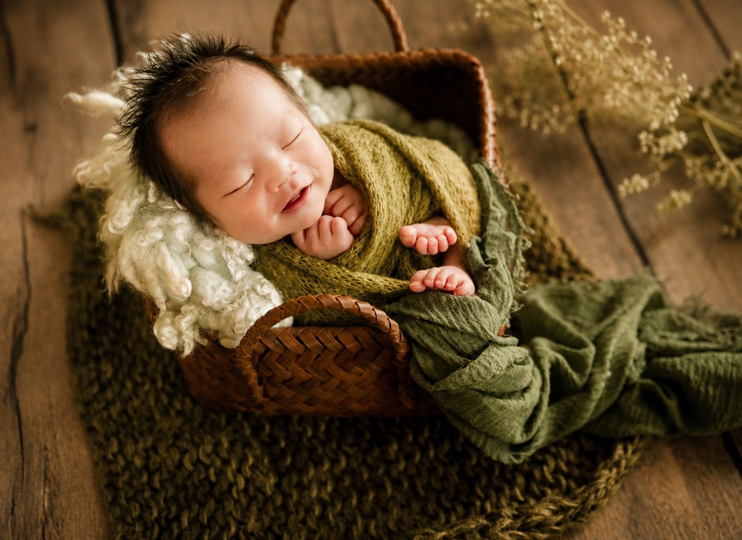 Smiling newborn baby wrapped in green swaddle sleeping peacefully in a wicker basket on a wooden floor.