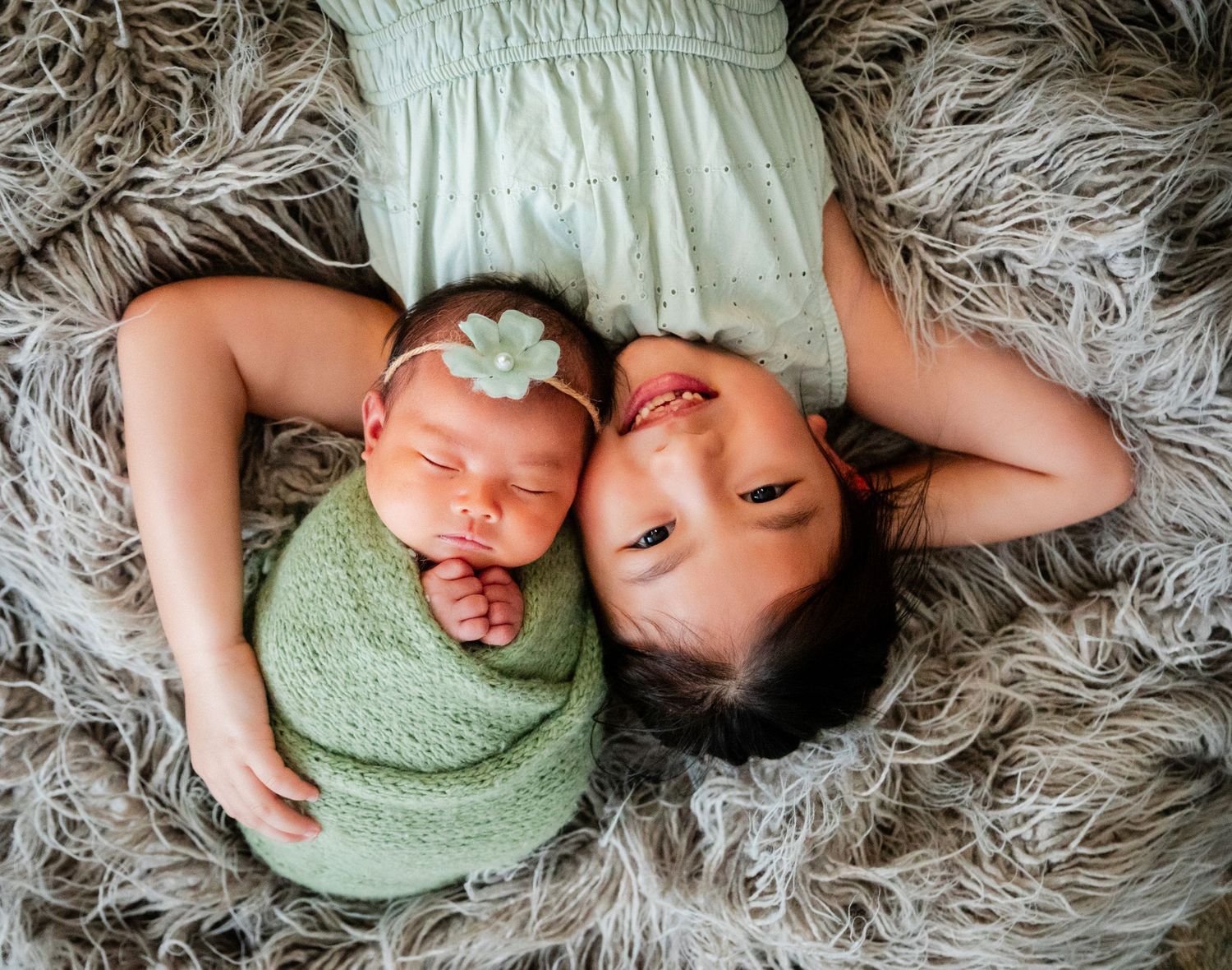 A young girl lies on a fluffy gray rug beside a swaddled newborn baby in green wrap with a floral headband.