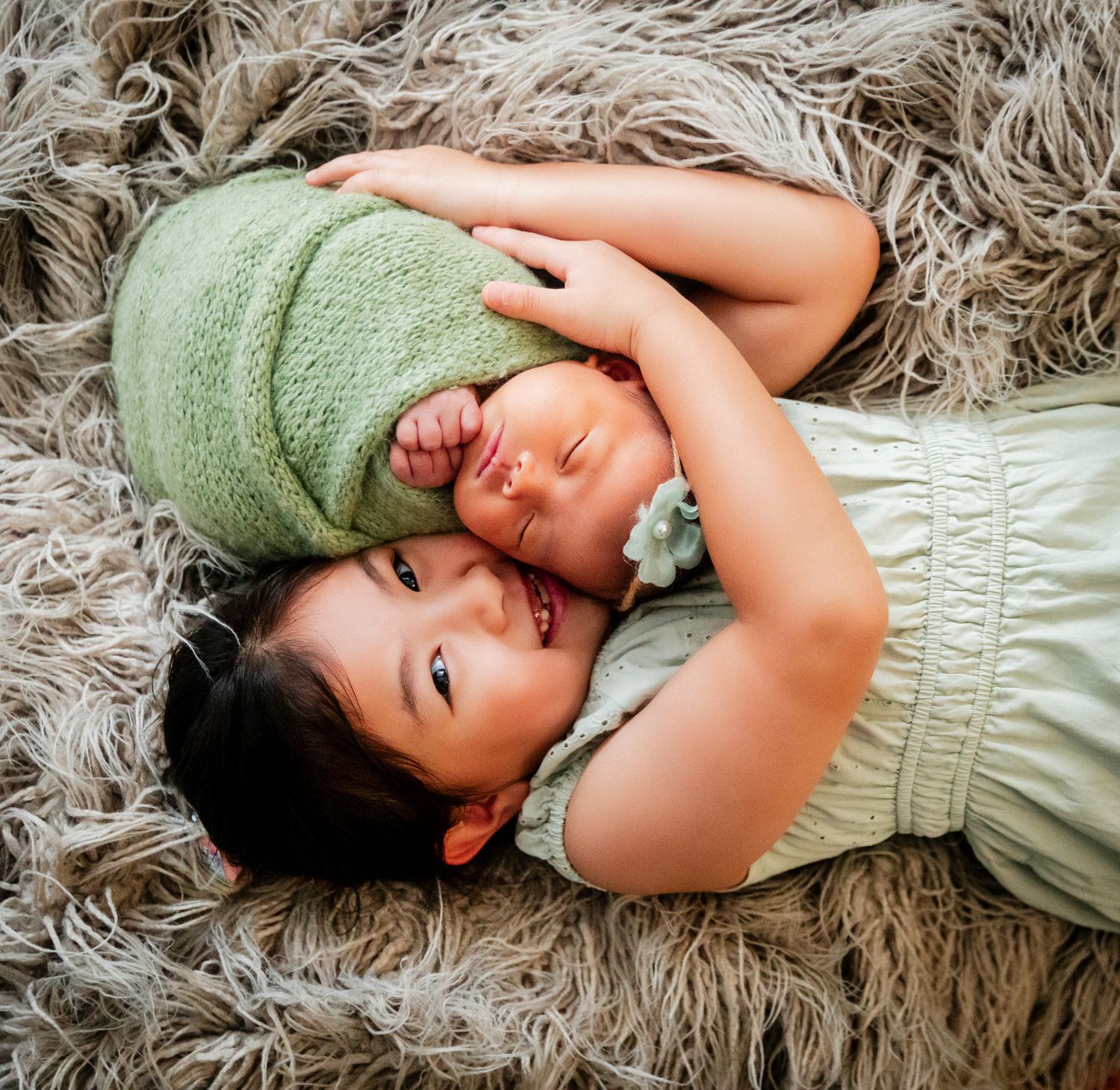 Young girl lying on fluffy rug cuddling newborn baby sibling wrapped in green knit blanket.