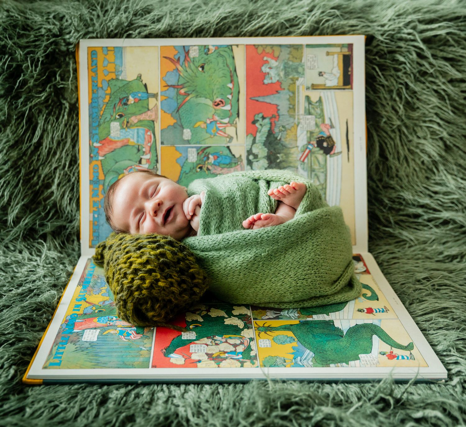 Smiling newborn baby wrapped in green knit blanket posed in open storybook on green fur backdrop.