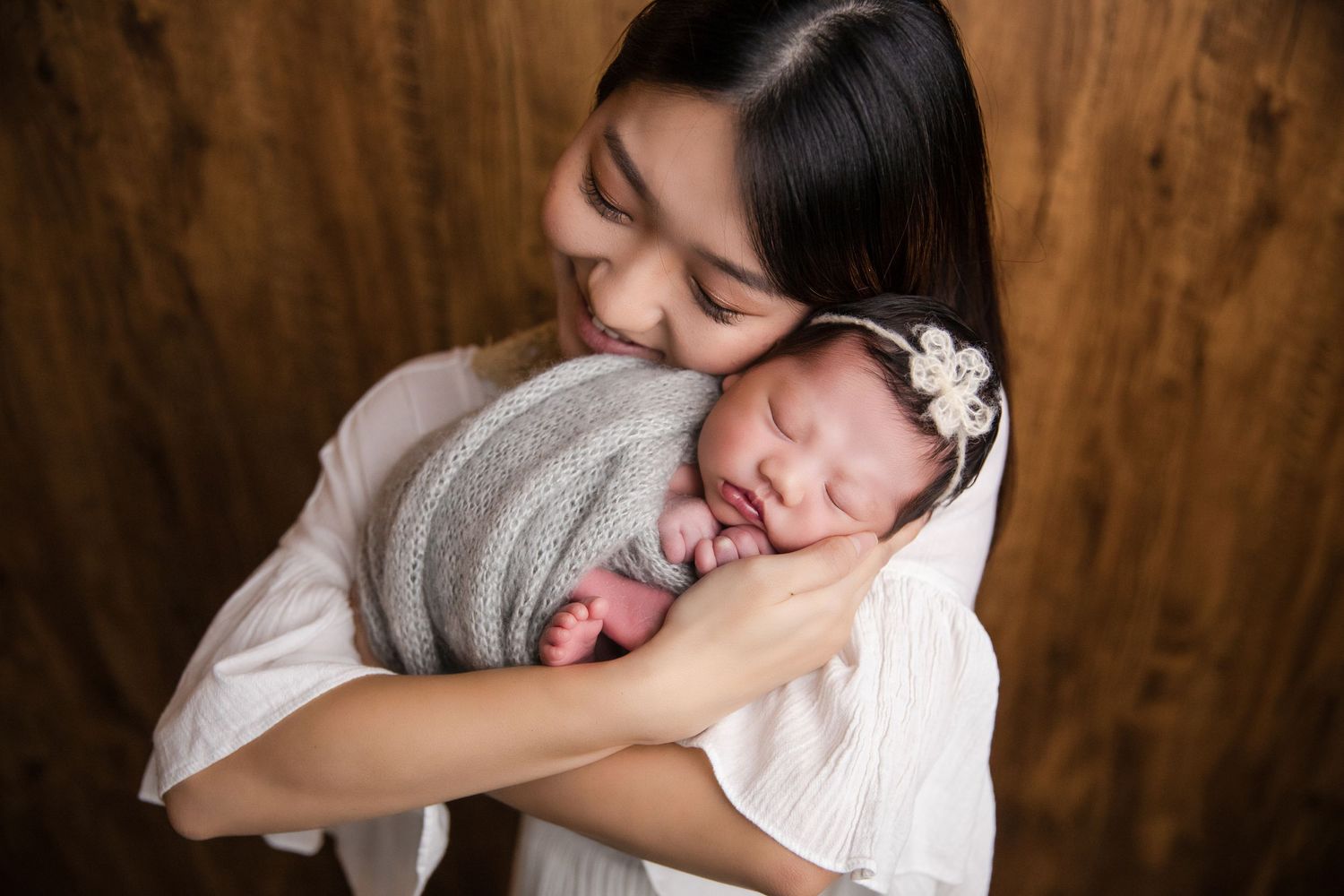 A mother tenderly holds her sleeping newborn wrapped in gray, both sharing a peaceful moment against a wood backdrop.