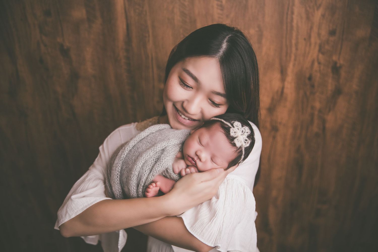 A smiling Asian mother tenderly holds her sleeping newborn baby wrapped in gray against a wooden background.