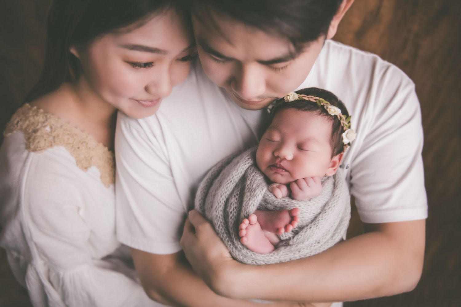 Young Asian parents tenderly holding their sleeping newborn baby wrapped in gray, with a floral headband.