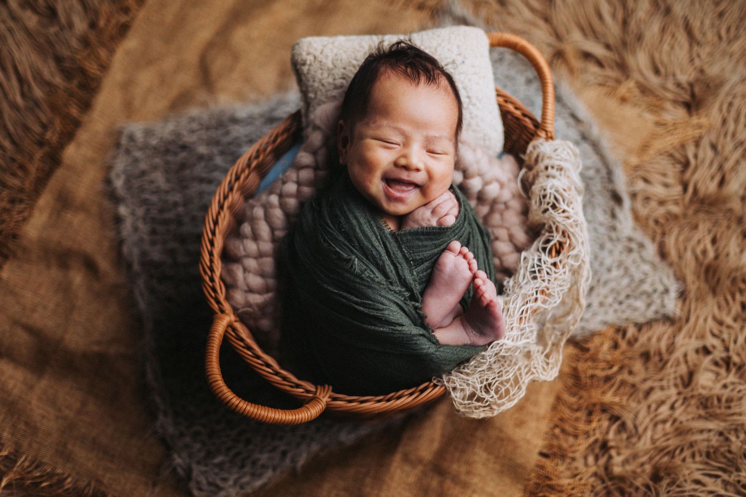 Smiling newborn baby wrapped in green swaddle, resting in a wicker basket with soft gray blankets.