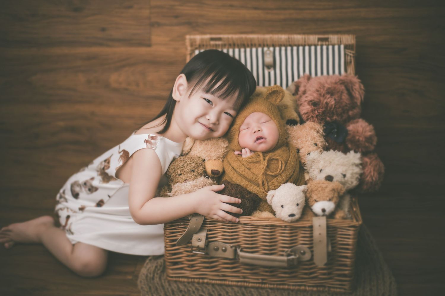 A smiling young girl poses with a sleeping newborn baby dressed in a lion costume inside a wicker basket with teddy bears.