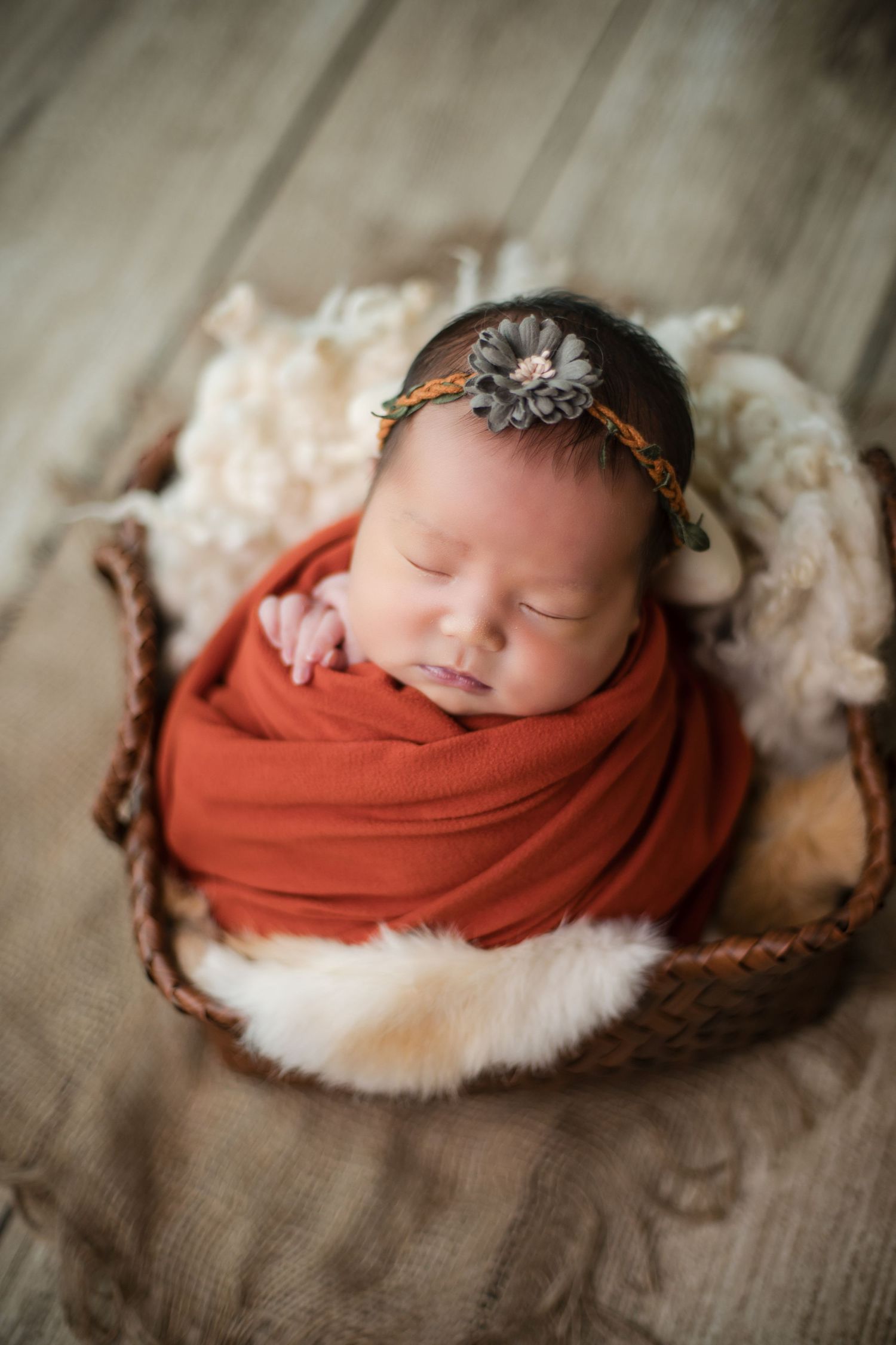 Newborn baby swaddled in rust orange wrap, wearing floral headband, sleeping peacefully in a wicker basket with fur lining.