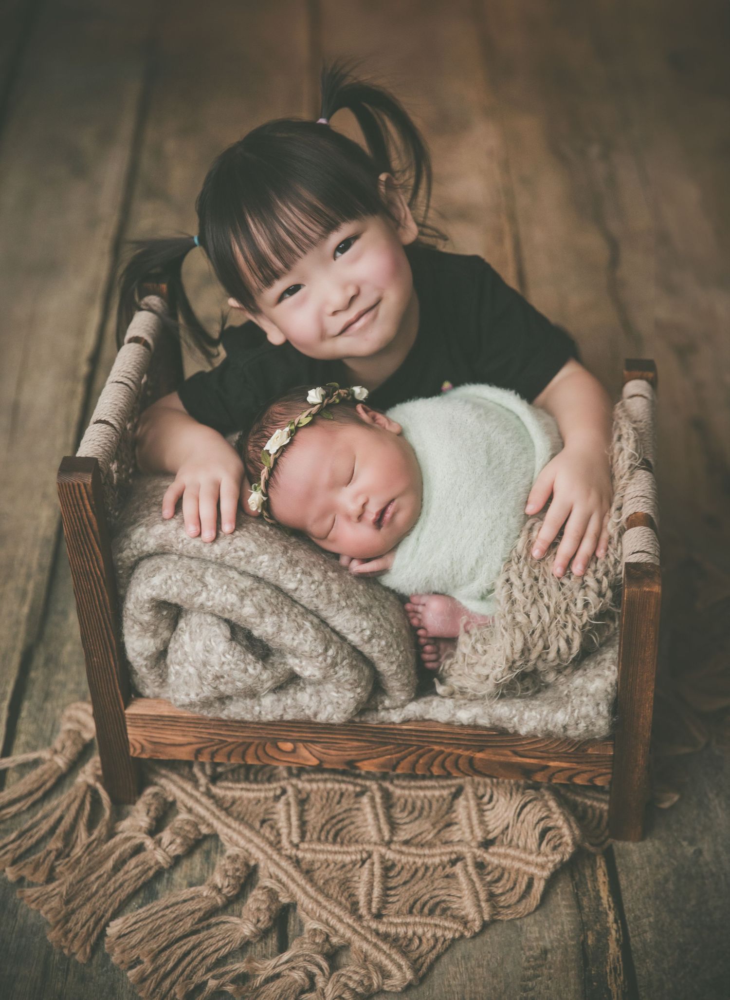 A young girl with pigtails smiles behind a sleeping newborn wrapped in white, lying in a rustic wooden mini bed.