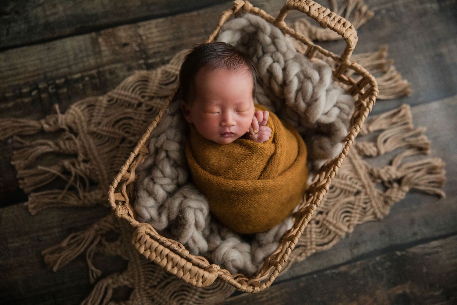 Newborn baby swaddled in mustard wrap sleeping in a wicker basket with chunky knit blanket on wood floor.
