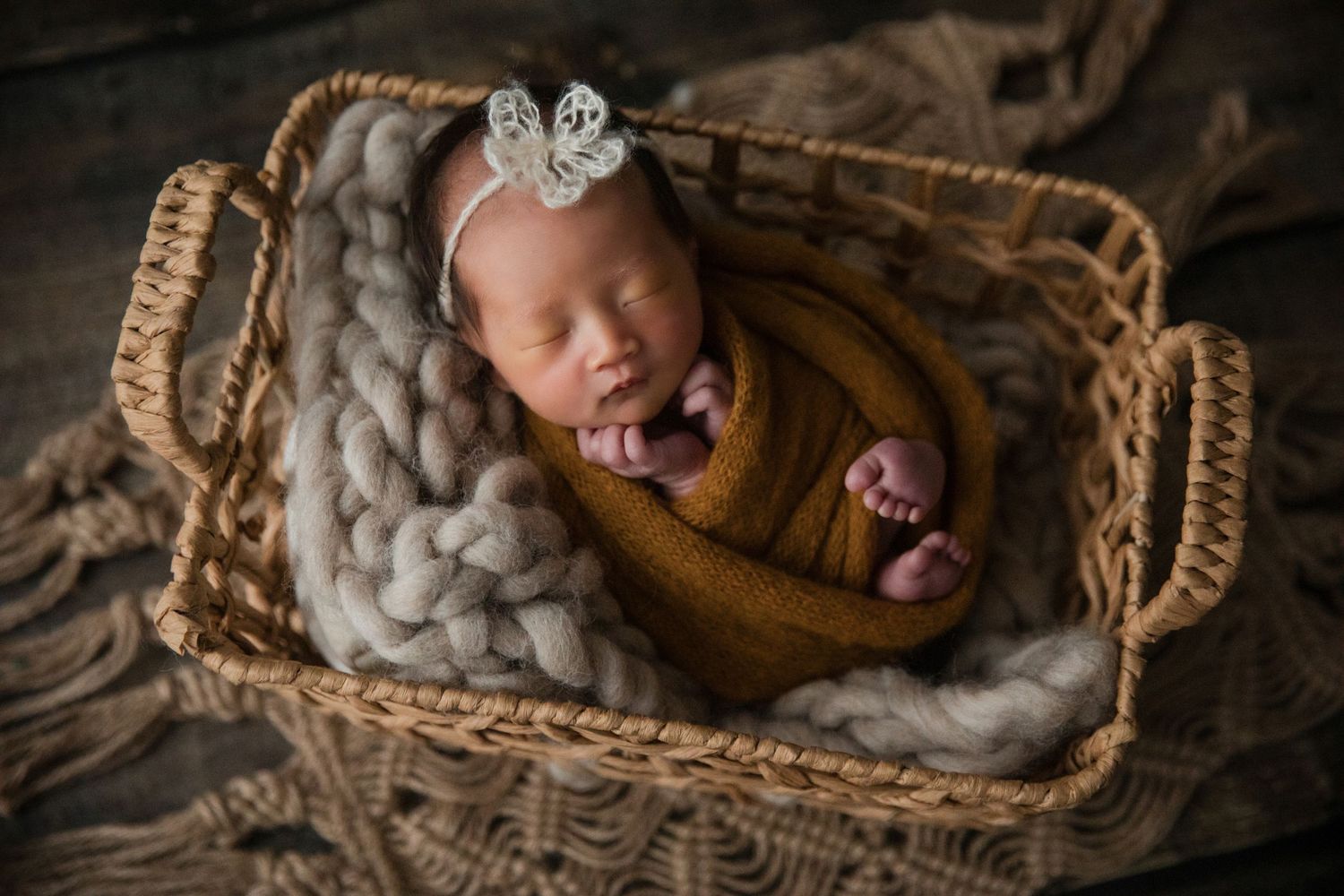 Newborn baby girl swaddled in mustard wrap, sleeping in wicker basket with chunky knit blanket and floral headband.