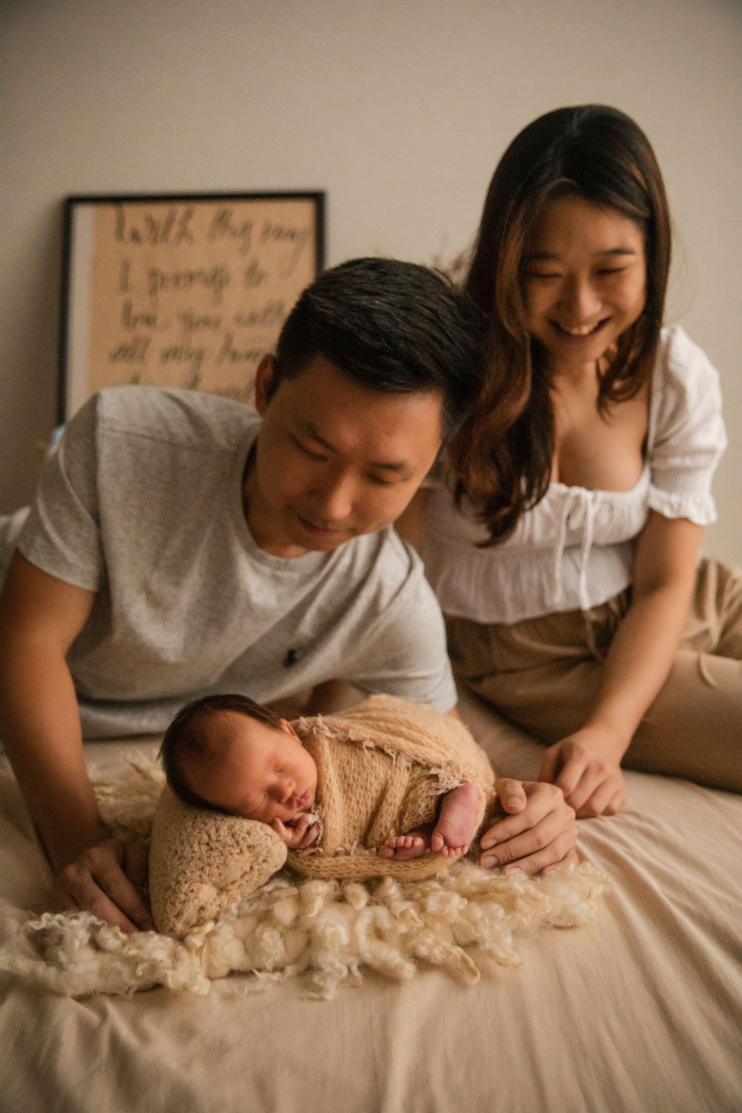 Happy Asian parents smiling at their sleeping newborn baby wrapped in a beige swaddle during a studio photoshoot.