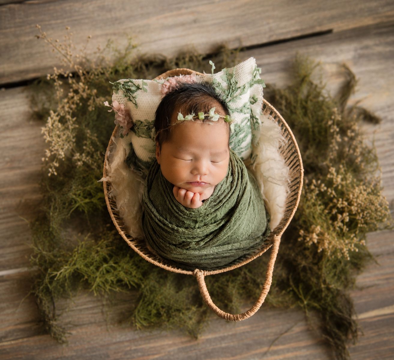 Newborn baby wrapped in green swaddle sleeping in wicker basket with floral headband and greenery.