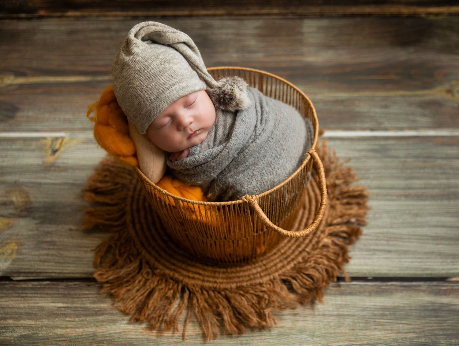 Newborn baby swaddled in gray wrap wearing knit hat sleeping peacefully in a rustic wooden bucket on burlap mat.