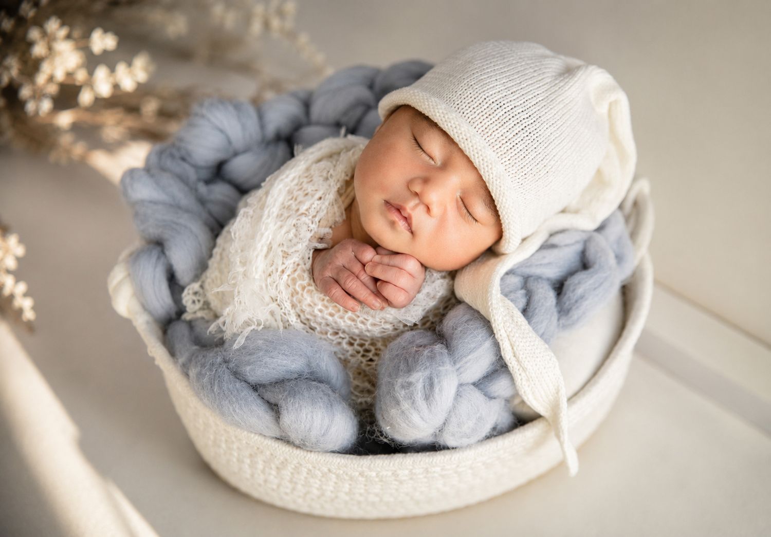 Sleeping newborn baby in white knit bonnet nestled in cozy blue yarn bowl for professional newborn photography.