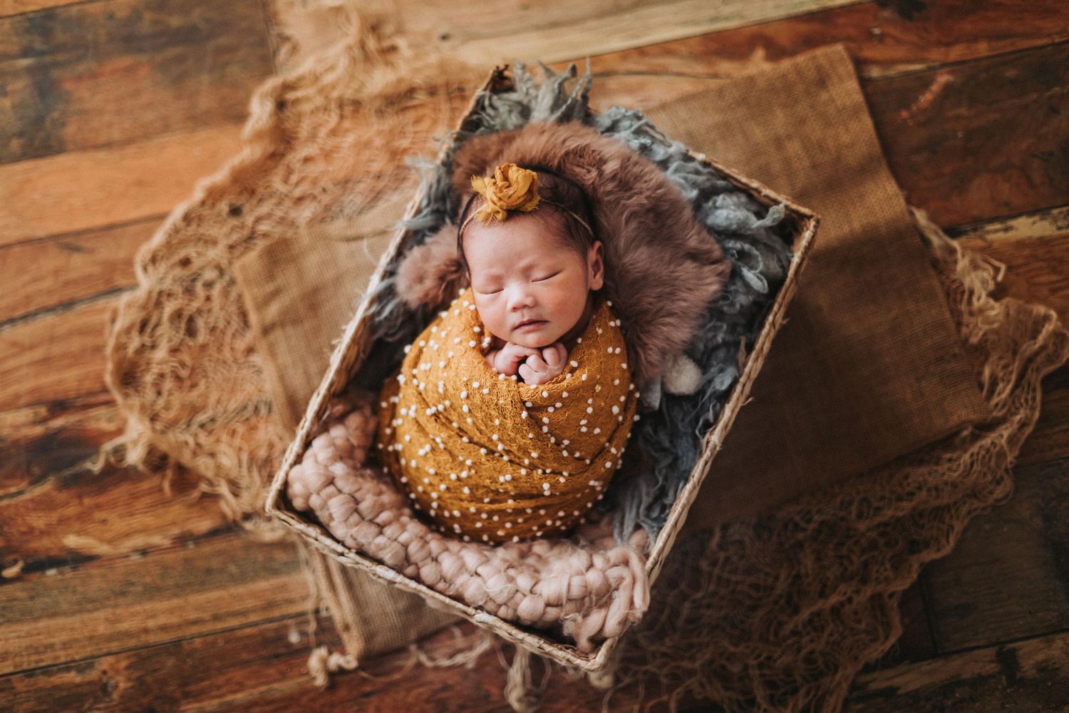 Newborn baby sleeping peacefully in a wooden box, wrapped in a mustard yellow knit swaddle with a matching bow headband.