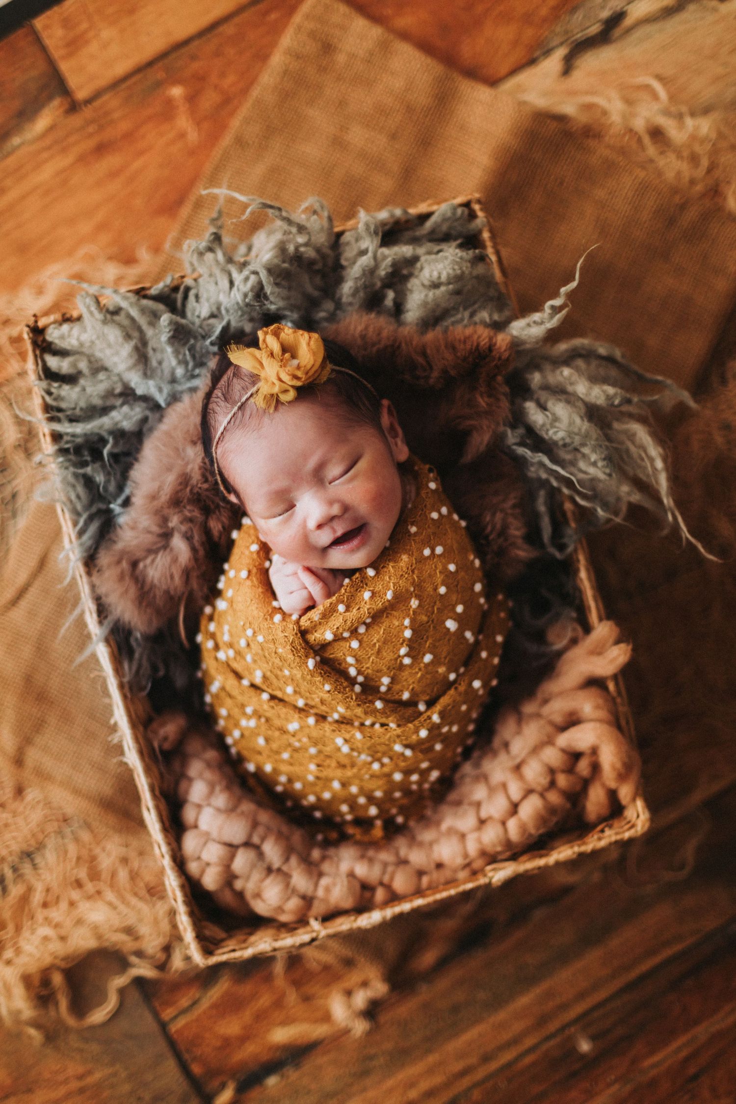 Newborn baby swaddled in mustard yellow wrap with floral headband, sleeping peacefully in a rustic wooden box.