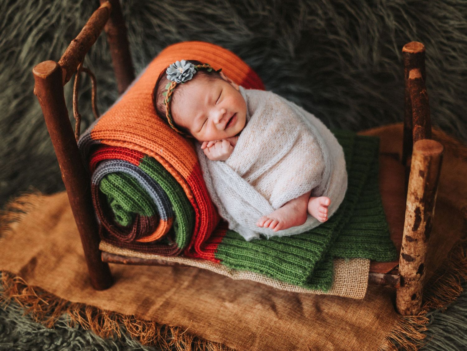 Newborn baby sleeping peacefully in a tiny rustic wooden bed with colorful knit blankets in a professional photo session.