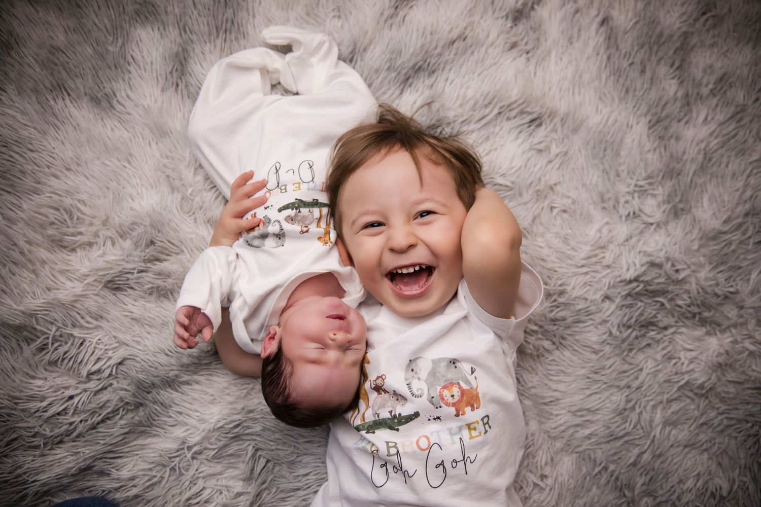 Two siblings, a laughing toddler and newborn baby, lying together on a fluffy gray fur rug in white onesies.