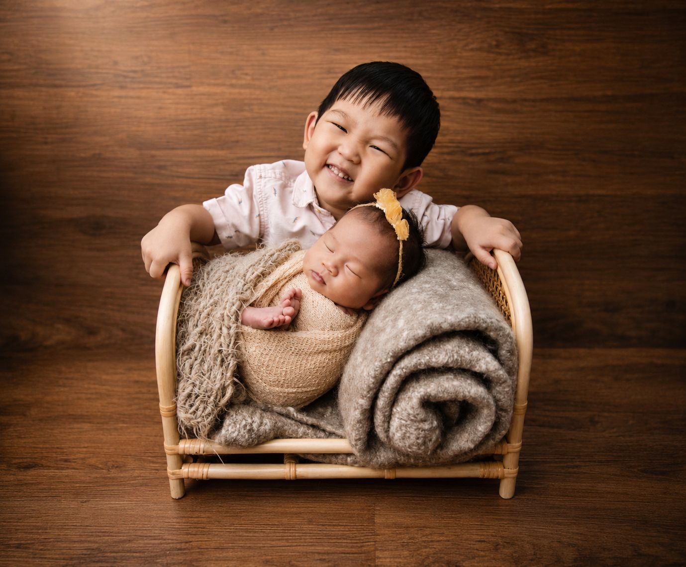 Smiling toddler boy poses with sleeping newborn baby sibling in a small wooden bed on hardwood floor.