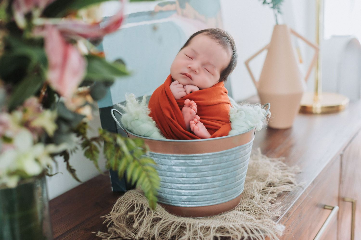 Newborn baby swaddled in orange wrap sleeping peacefully inside a rustic metal bucket.