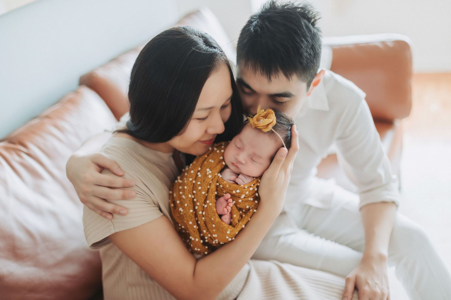 A couple tenderly kisses their sleeping newborn baby wrapped in a golden floral swaddle during a family portrait session.