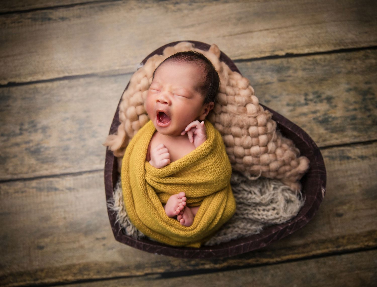 Newborn baby swaddled in yellow wrap, yawning in heart-shaped wooden bowl with knit blanket on rustic wood floor.