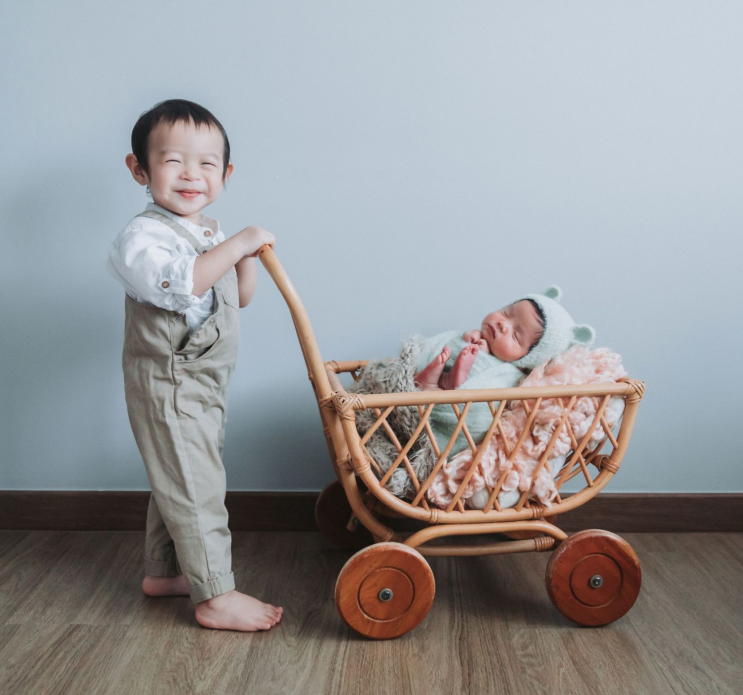 Smiling toddler boy pushes a wicker baby carriage with a sleeping newborn inside, against a light gray wall.
