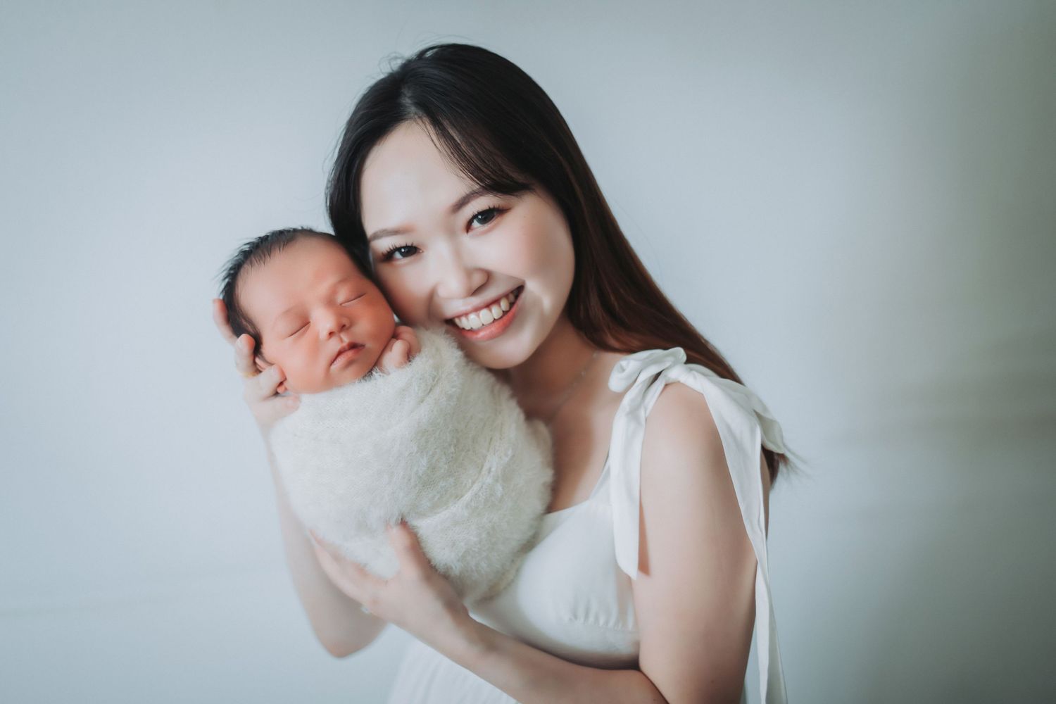 Smiling young Asian mother holding her sleeping newborn baby wrapped in a white fluffy blanket.