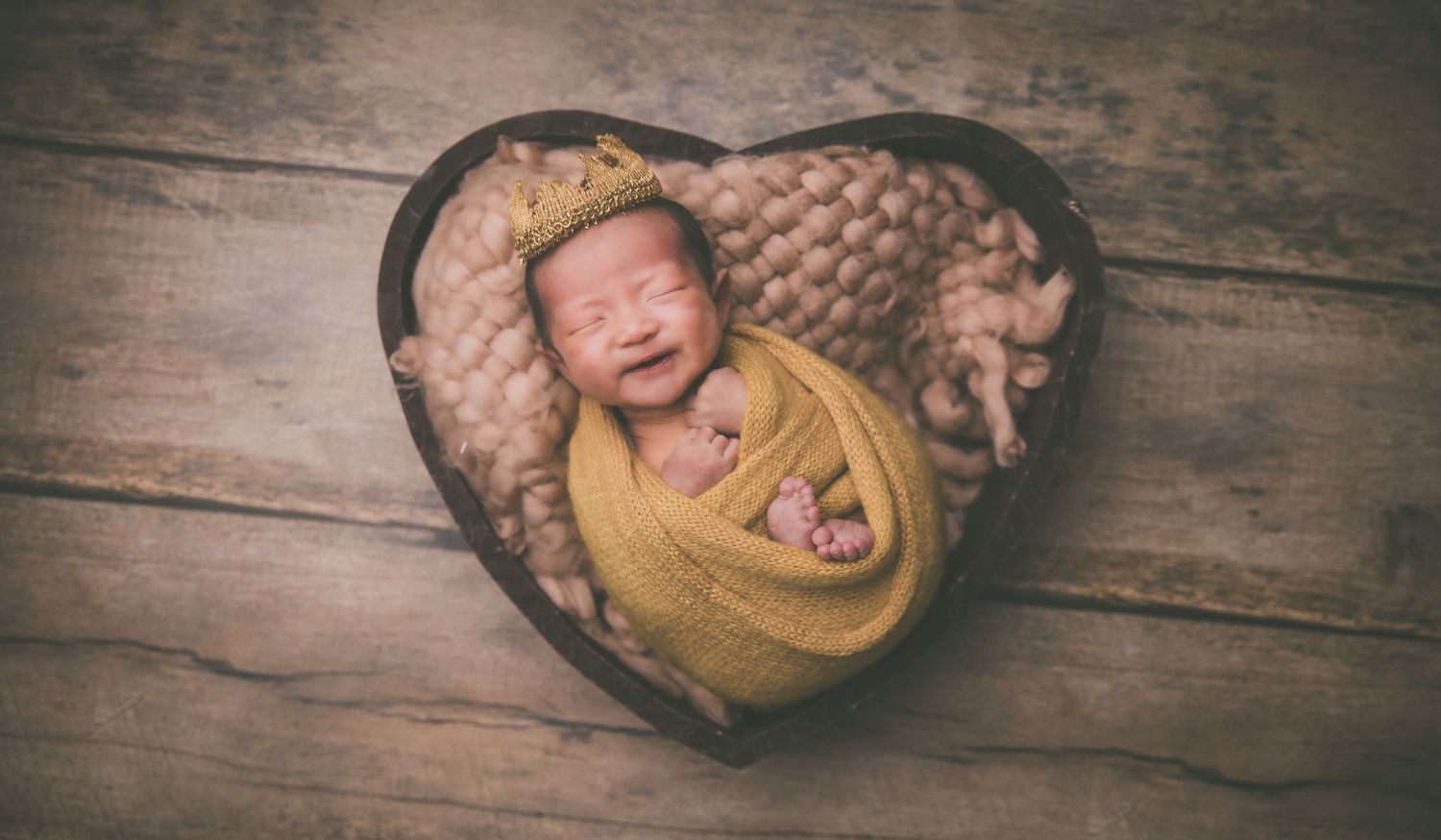 Smiling newborn baby swaddled in yellow wrap wearing tiny crown nestled in heart-shaped wooden bowl with soft blanket.
