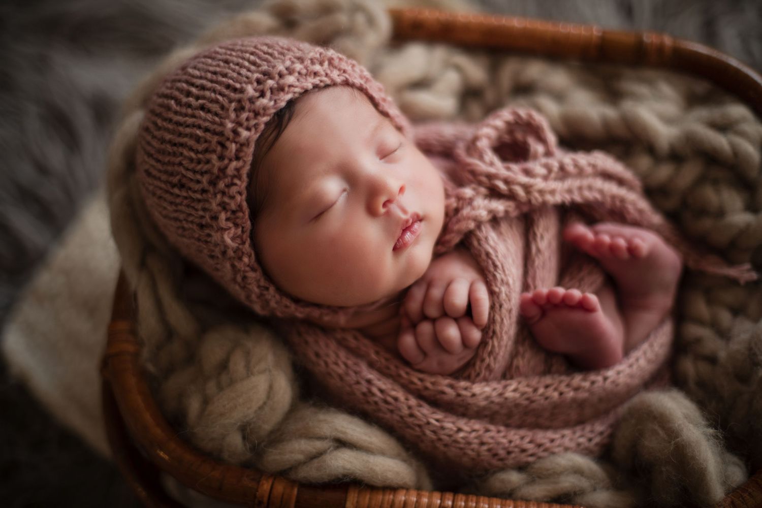 Newborn baby sleeping peacefully in a basket, wrapped in a pink knit blanket and bonnet.