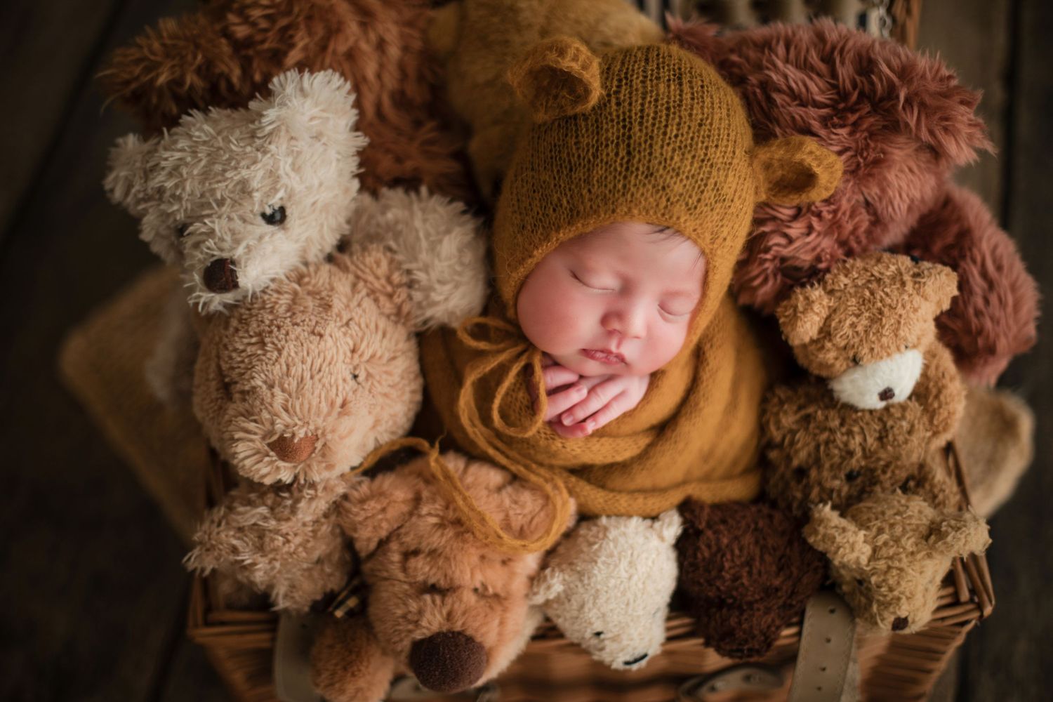 Sleeping newborn baby in mustard bear hat nestled among teddy bears in a wicker basket.