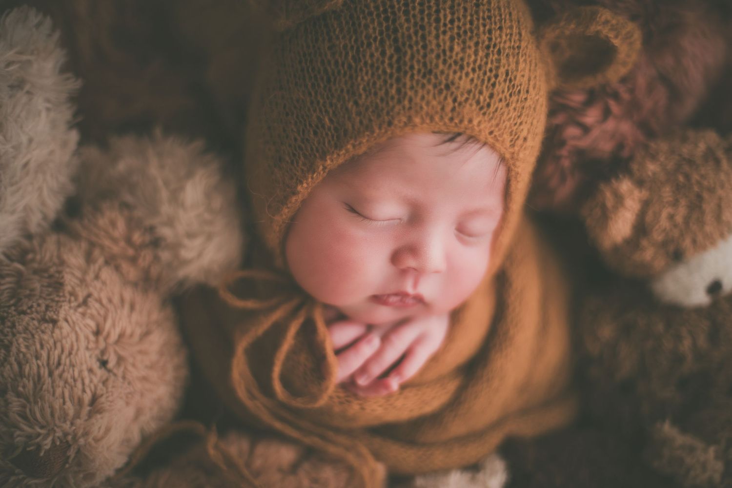 Sleeping newborn baby wearing a mustard knit bonnet surrounded by soft teddy bears in a cozy nest.