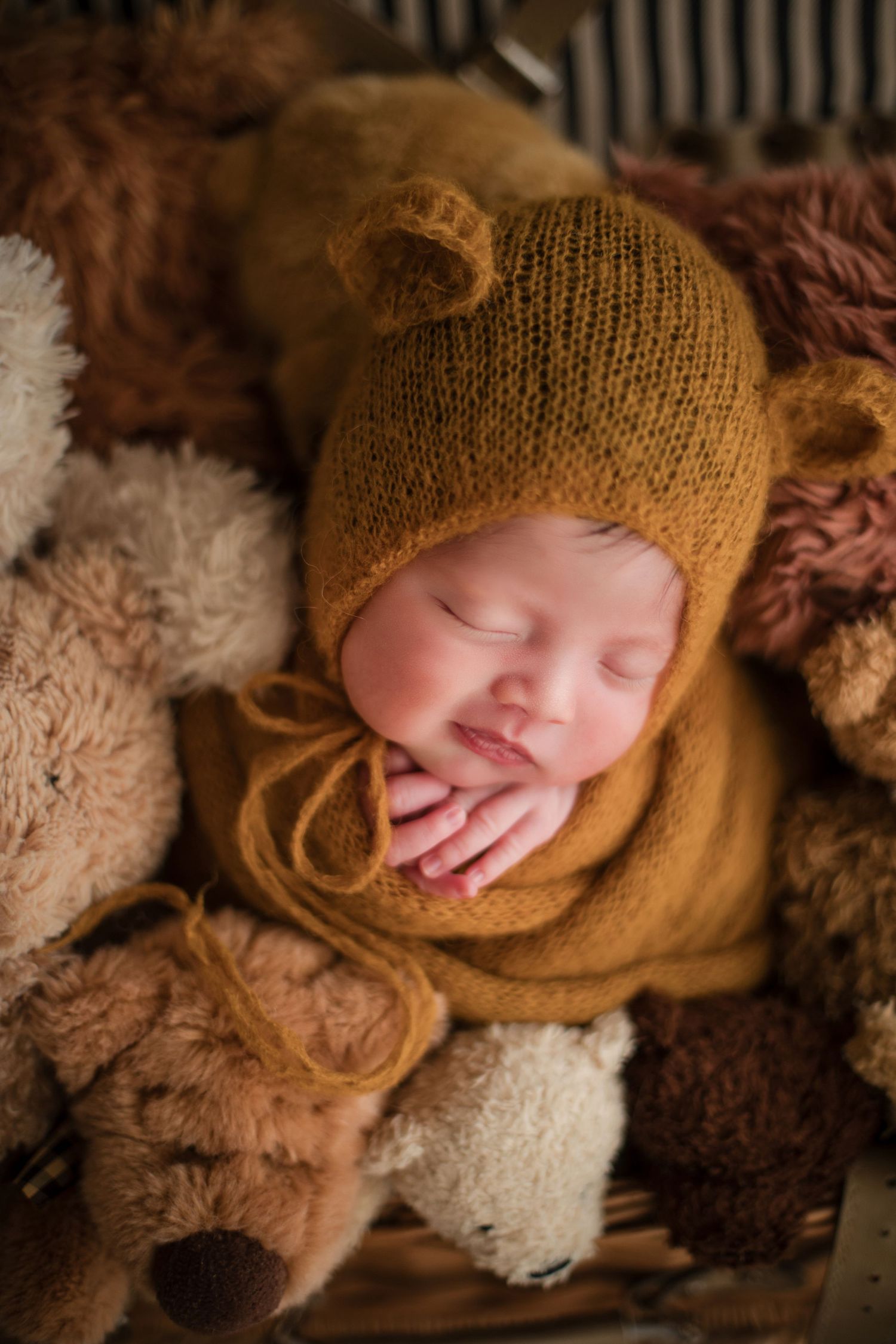 Sleeping newborn baby in mustard knit bear hat and wrap, nestled among teddy bears in a cozy basket.