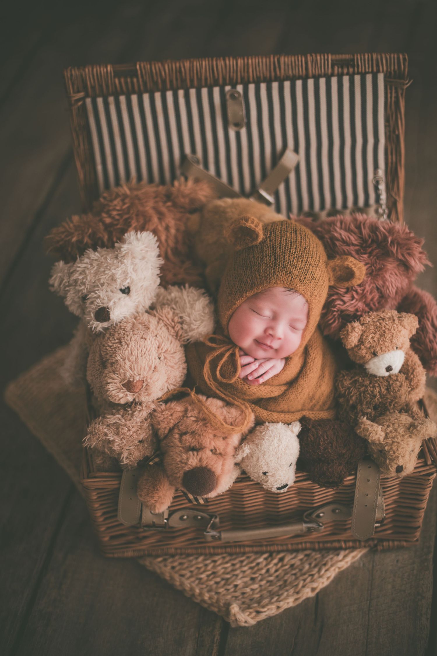 Newborn baby sleeping in wicker basket surrounded by teddy bears, wearing brown knit bonnet.
