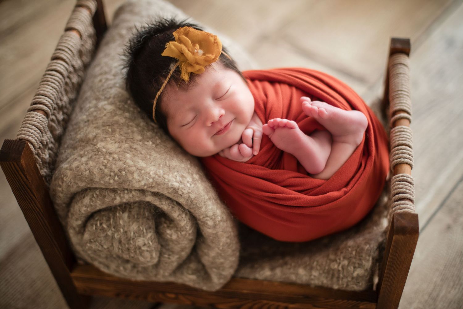 Newborn baby swaddled in orange wrap with gold flower headband sleeping peacefully in a tiny wooden bed.