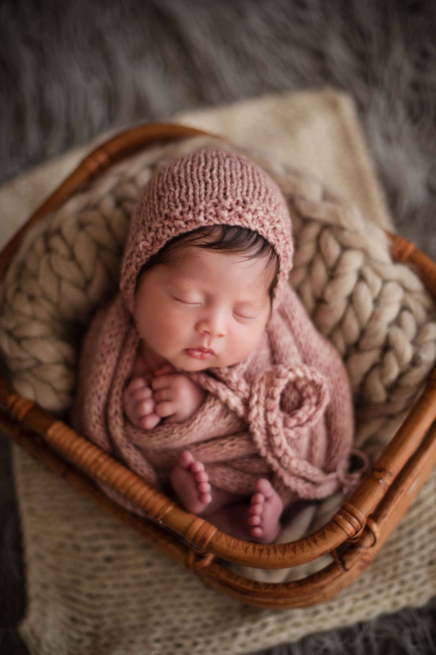 Newborn baby sleeping peacefully in a wicker basket, wrapped in a soft pink knit blanket and matching bonnet hat.