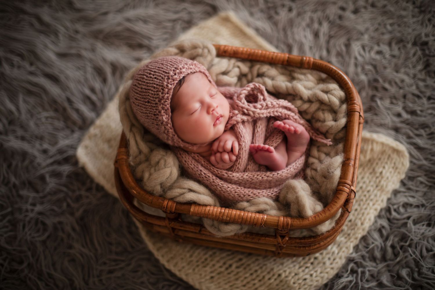 Newborn baby sleeping peacefully in a wicker basket, wrapped in pink knit blanket and bonnet on gray fur.