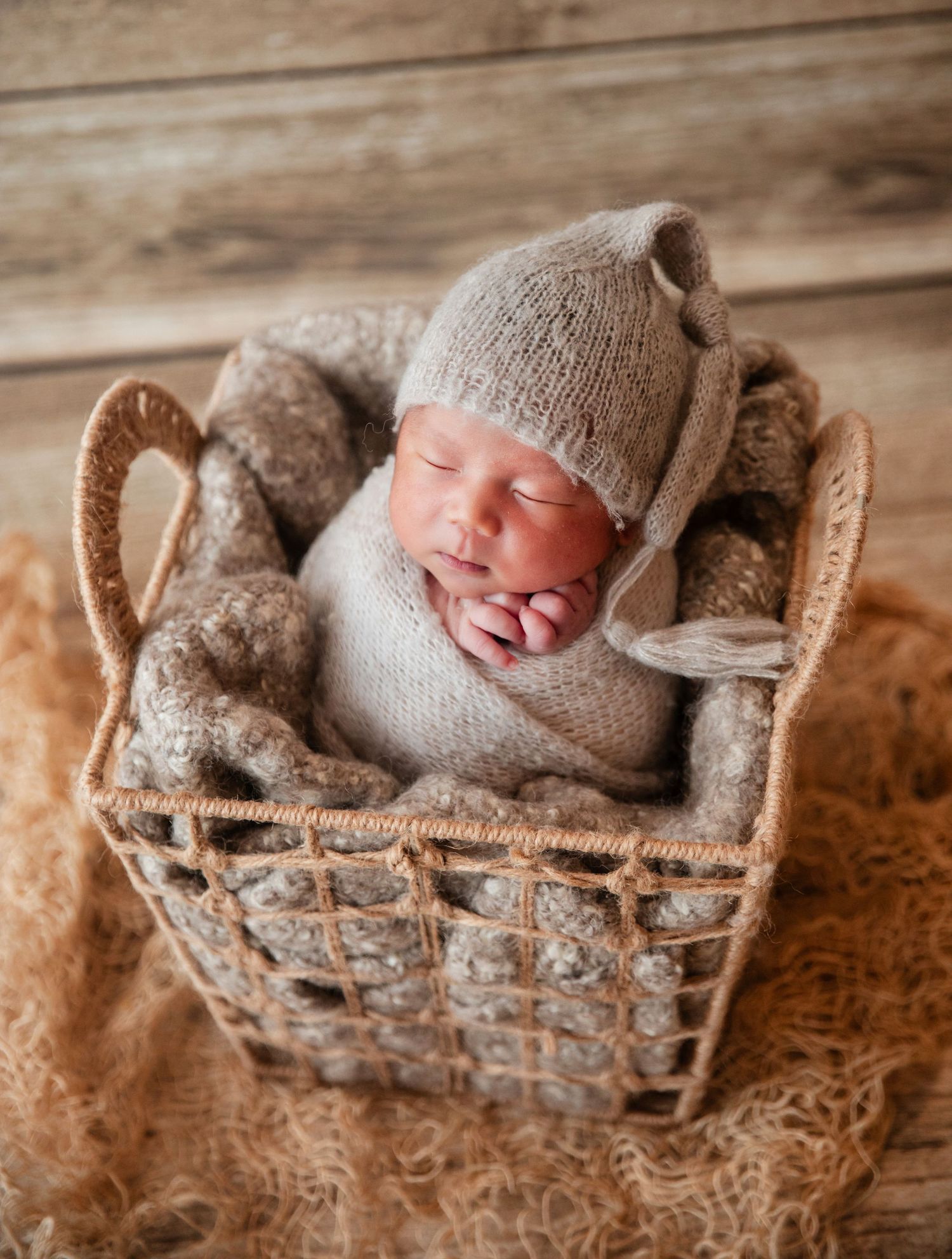 Sleeping newborn baby wrapped in beige knit blanket wearing a cozy knit hat, nestled in a wicker basket.