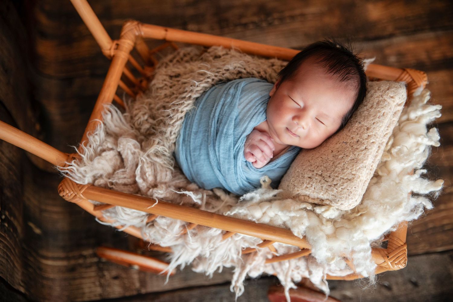 Newborn baby swaddled in blue wrap sleeping peacefully in a rustic wooden basket with fluffy blanket.
