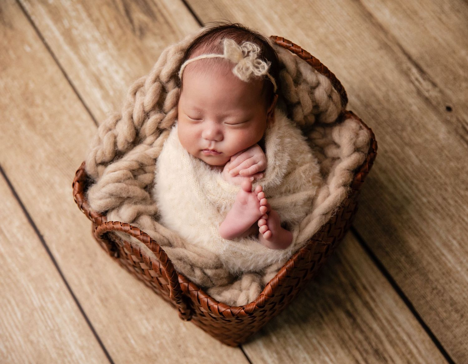 Sleeping newborn baby wrapped in beige knit blanket inside a wicker basket with a floral headband.