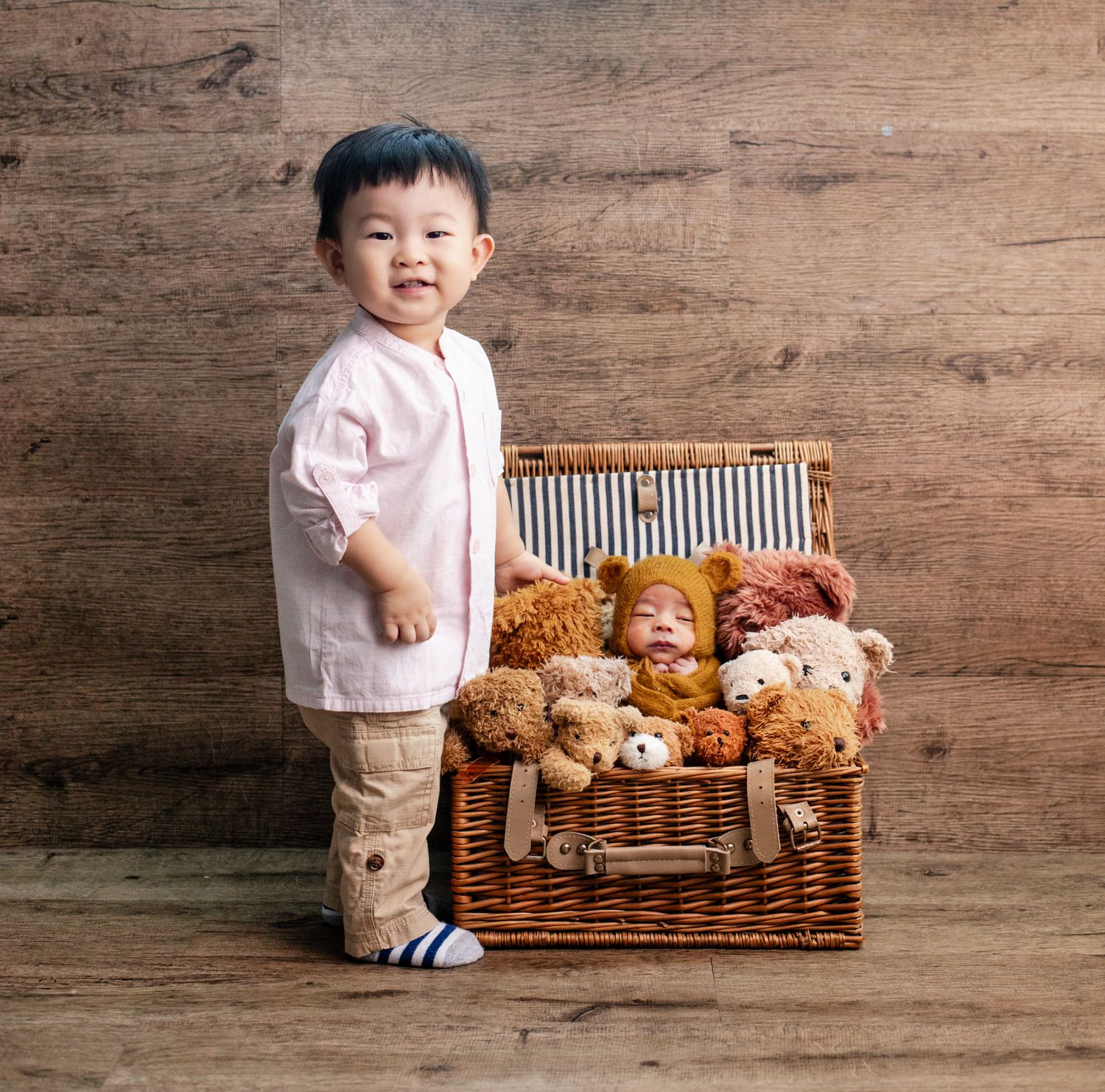 A smiling toddler stands beside a wicker basket filled with teddy bears and a sleeping newborn baby.